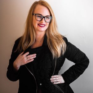 Melanie smiling. Wearing a black jacket, black glasses and red lipstick. strawberry blonde hair